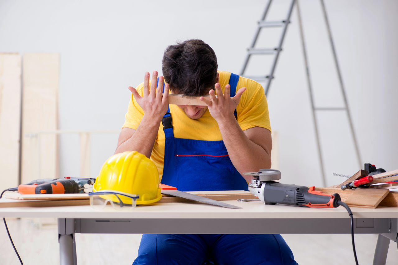 Bouwvakker in werkkleding zit aan een werkbank met gereedschap en houdt zijn hoofd vast uit frustratie tijdens het werk.