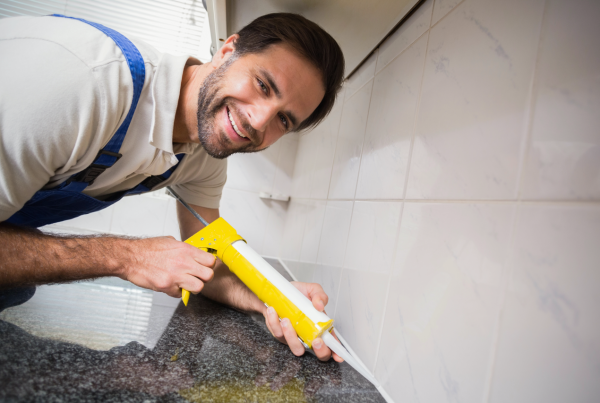 Smiling worker in overalls uses a caulking gun to neatly seal joints along a kitchen countertop.