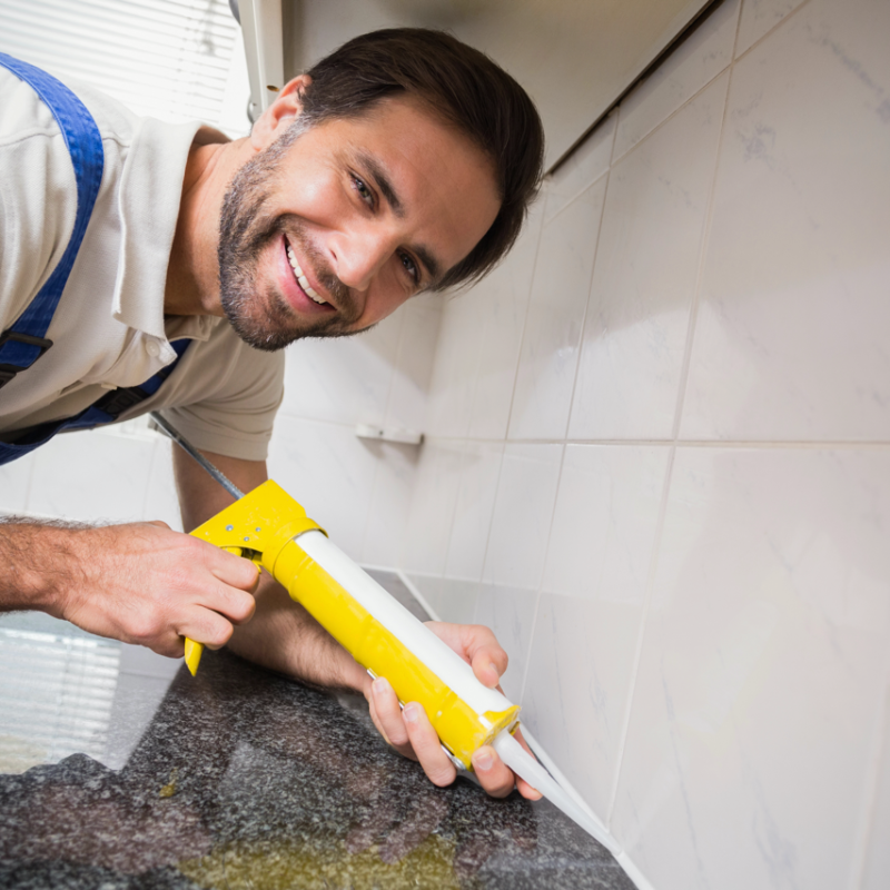 Smiling worker in overalls uses a caulking gun to neatly seal joints along a kitchen countertop.