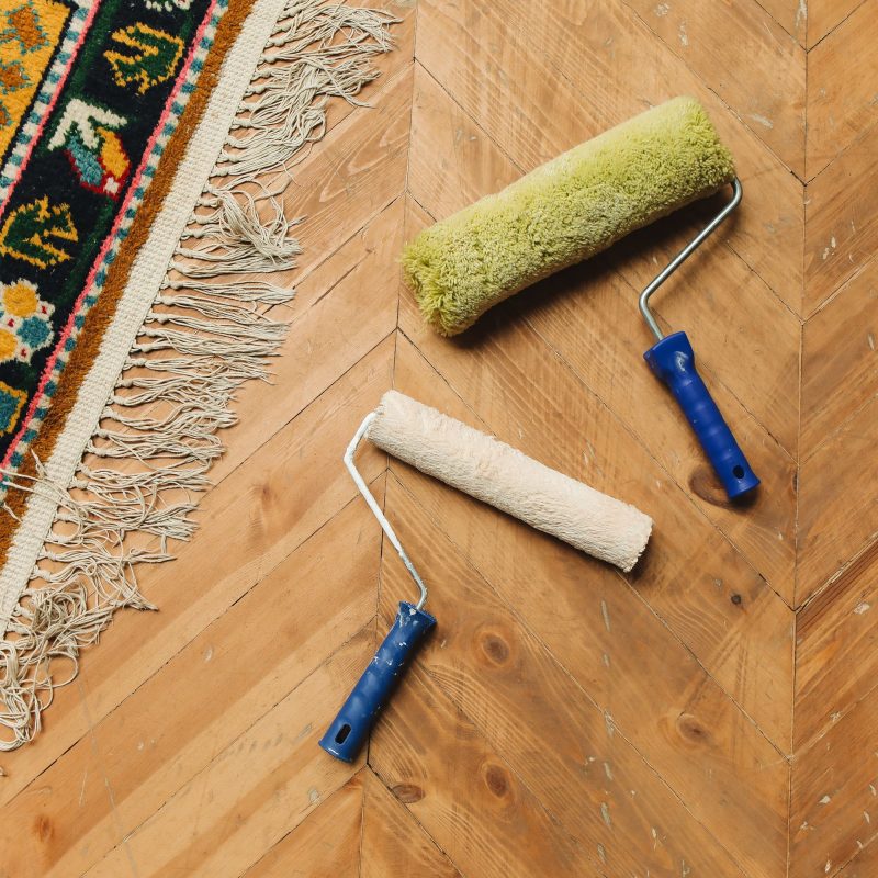 Een houten visgraatvloer met twee verfrollers erop. Aan de linkerkant ligt een kleurrijk vloerkleed met franjes.