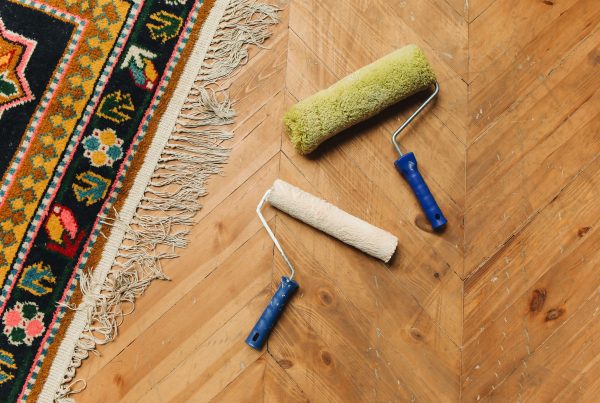 A wooden herringbone floor with two paint rollers placed on it. A colorful patterned rug with fringes is visible on the left side.