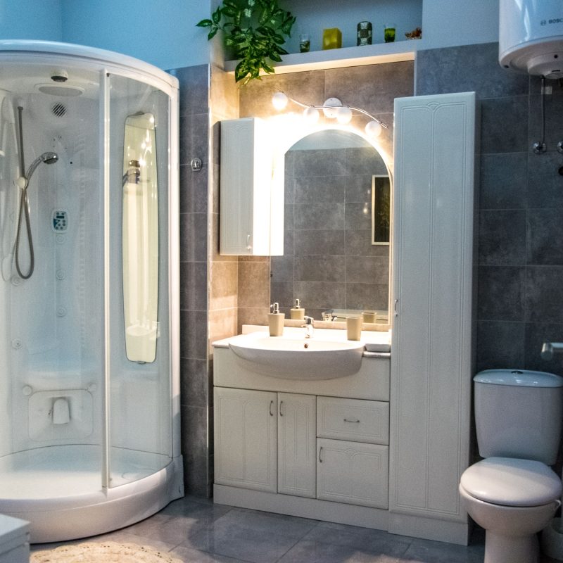 A bright and functional bathroom featuring a modern corner shower, a spacious vanity with a large mirror, practical storage cabinets, and neutral grey tiles. The clean design creates a comfortable and timeless atmosphere.