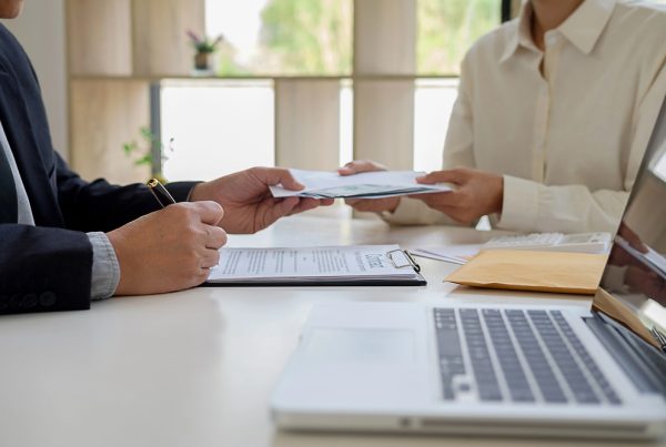 Two people at a table exchanging documents during a financial or advisory conversation, with a laptop, a clipboard with papers and an envelope on the table – an atmosphere of discussion about financing or a subsidy application.