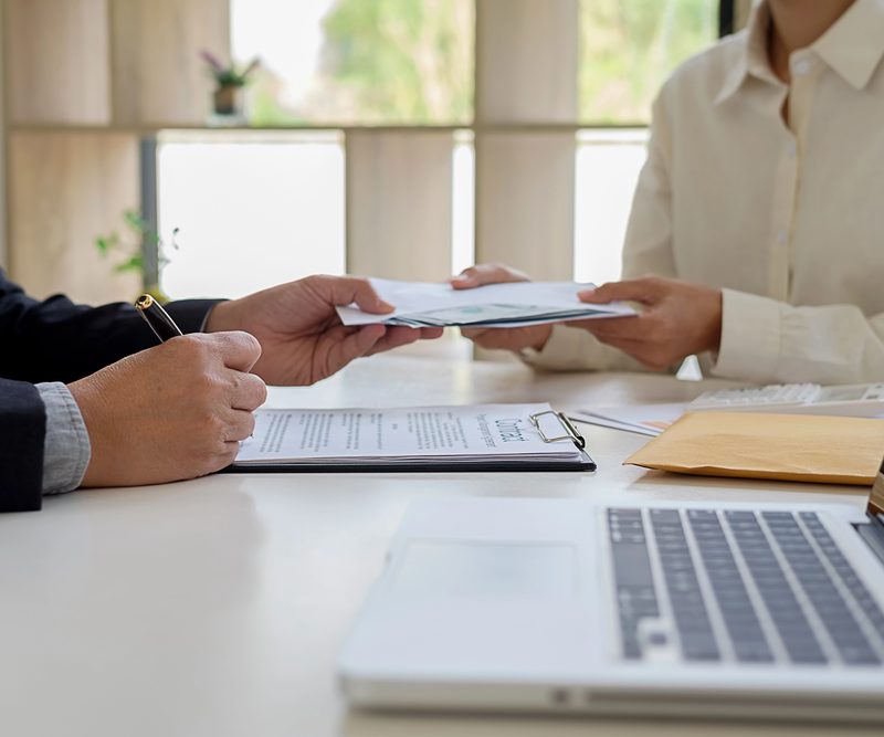 Two people at a table exchanging documents during a financial or advisory conversation, with a laptop, a clipboard with papers and an envelope on the table – an atmosphere of discussion about financing or a subsidy application.