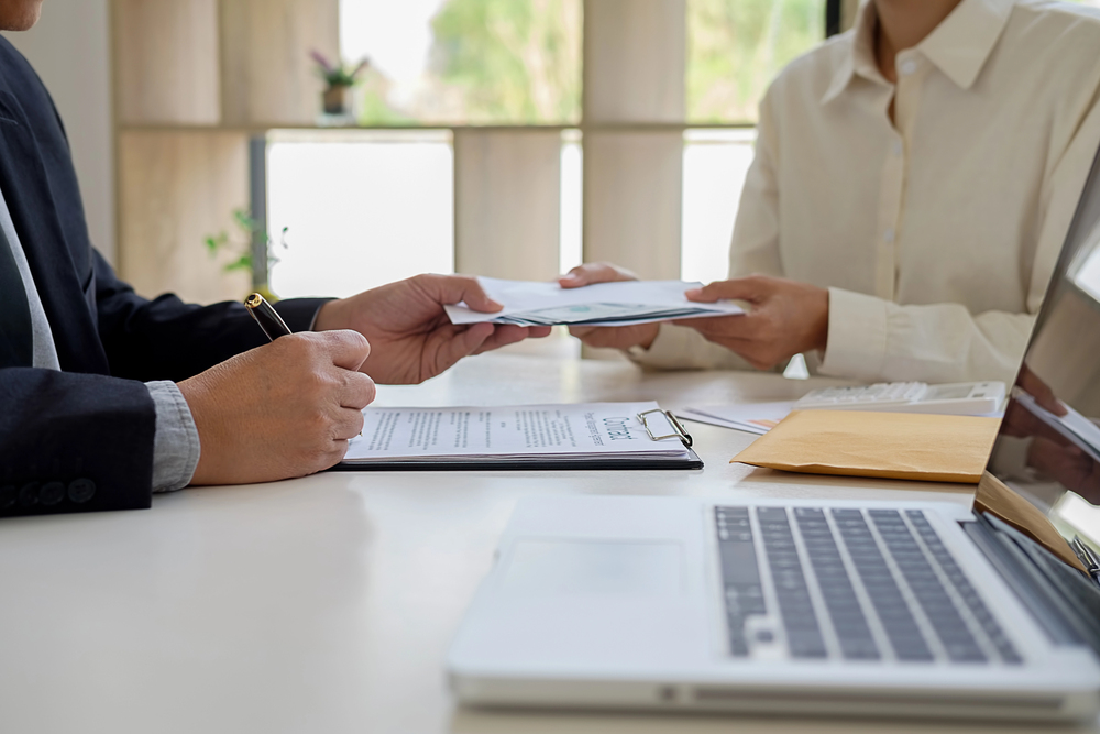 Two people at a table exchanging documents during a financial or advisory conversation, with a laptop, a clipboard with papers and an envelope on the table – an atmosphere of discussion about financing or a subsidy application.