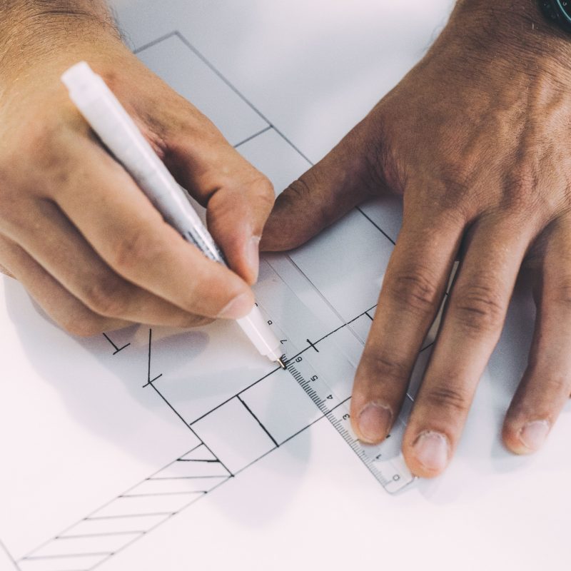 Op de foto zie je iemand die met een liniaal en een pen een technische tekening of bouwplan maakt.